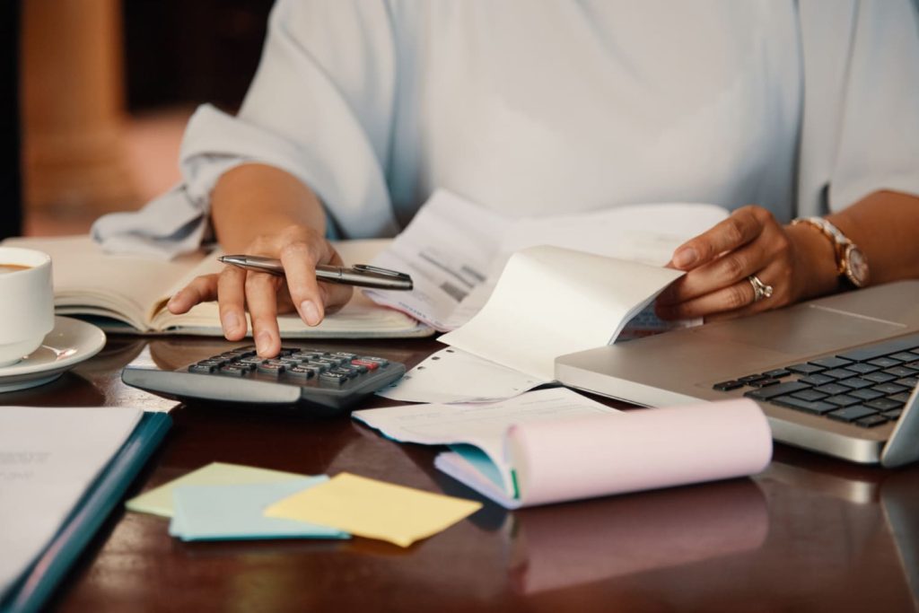 mulher usando calculadora com vários papéis e um notebook a sua volta